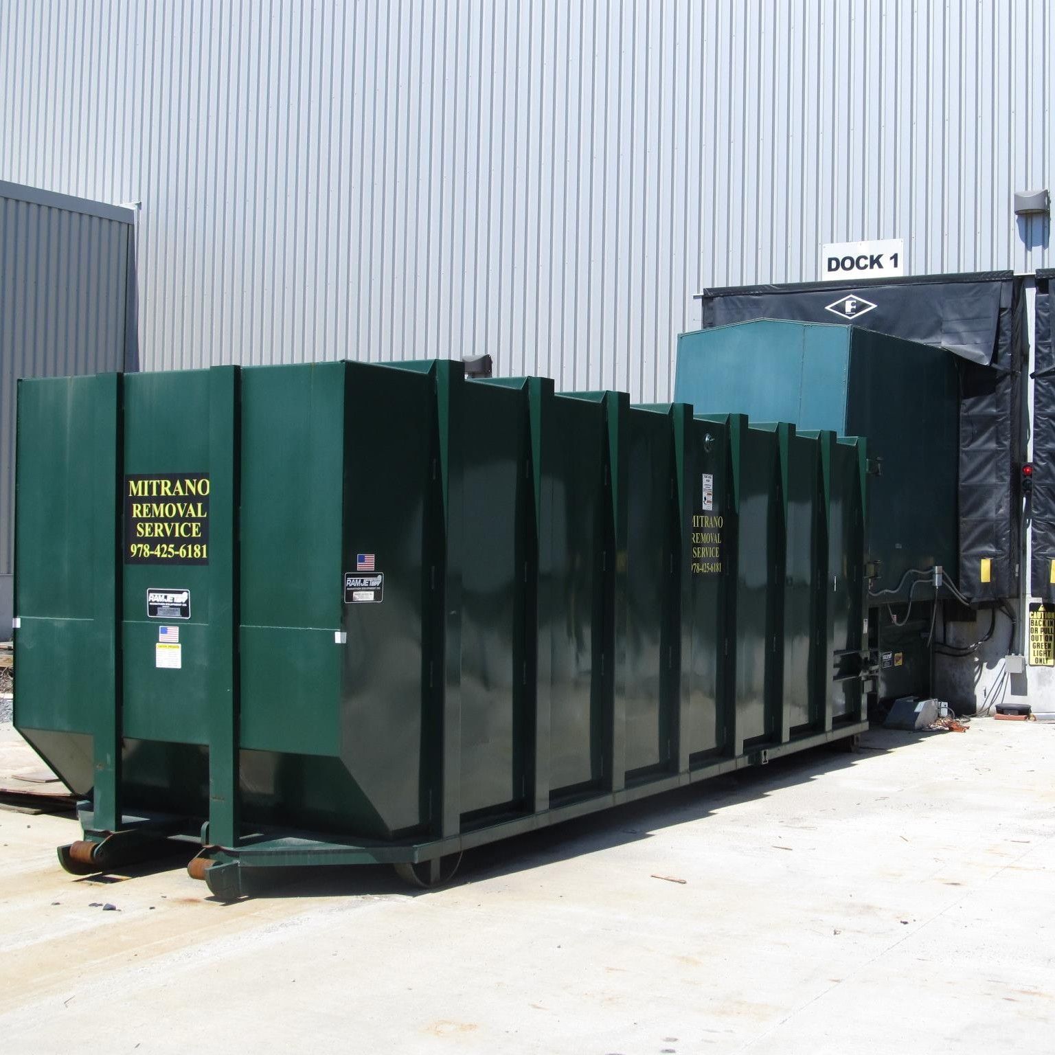 Dark green industrial waste compactor at a loading dock.
