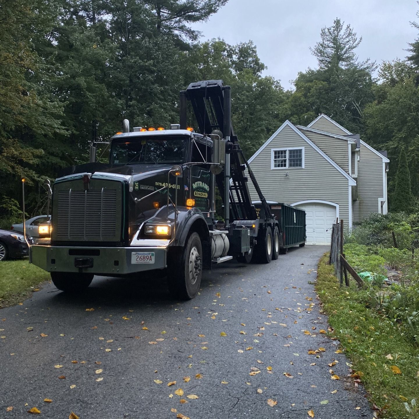 Black garbage truck with a dumpster parked in a driveway in front of a house.