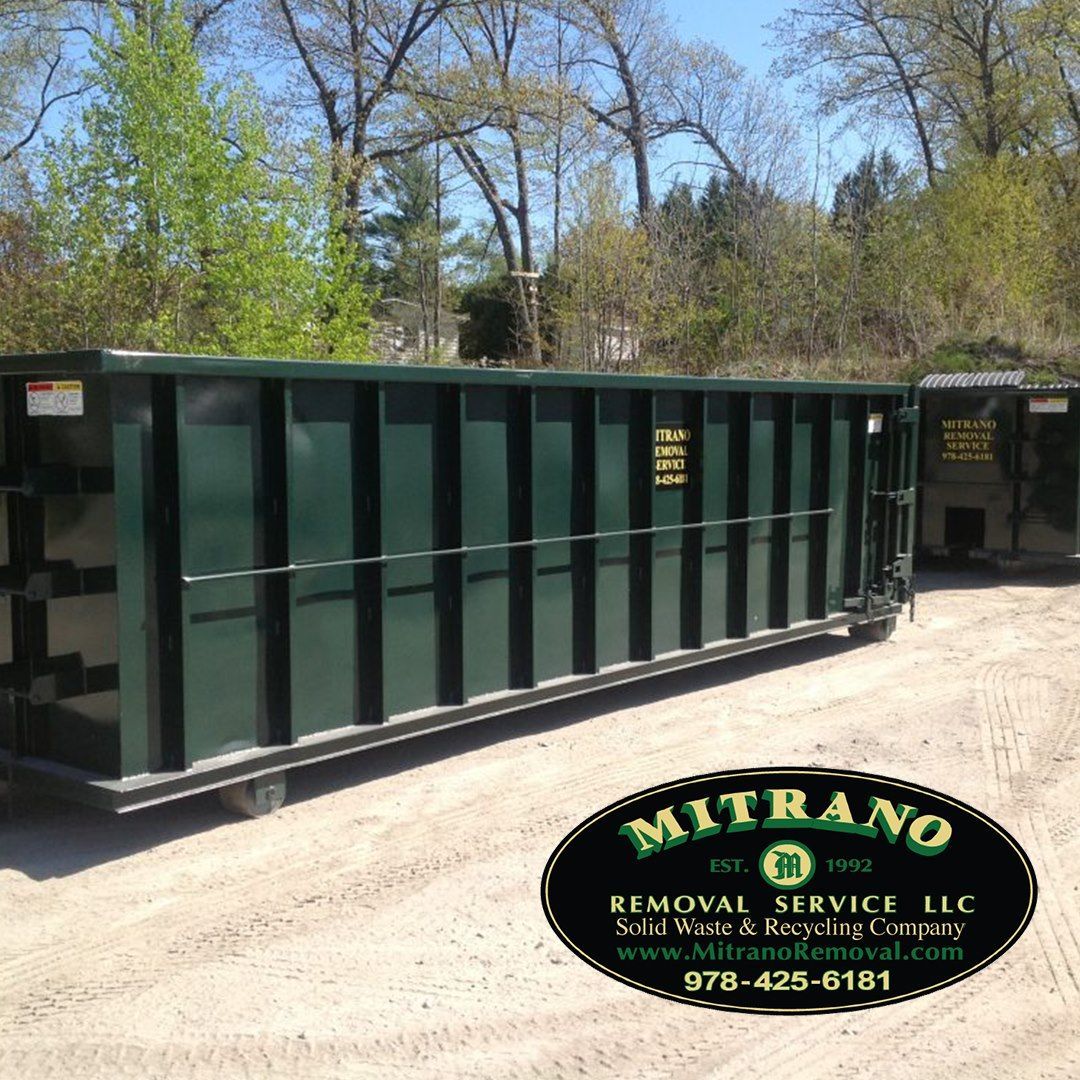 Green dumpster in a dirt lot, with trees in background. Mitrano Removal Service logo at the bottom.