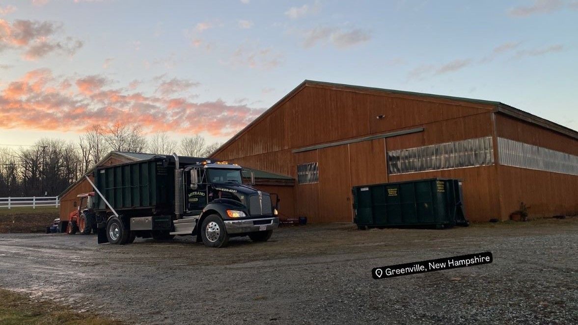 Dark dump truck beside a rusty warehouse; early morning sky, gravel lot.