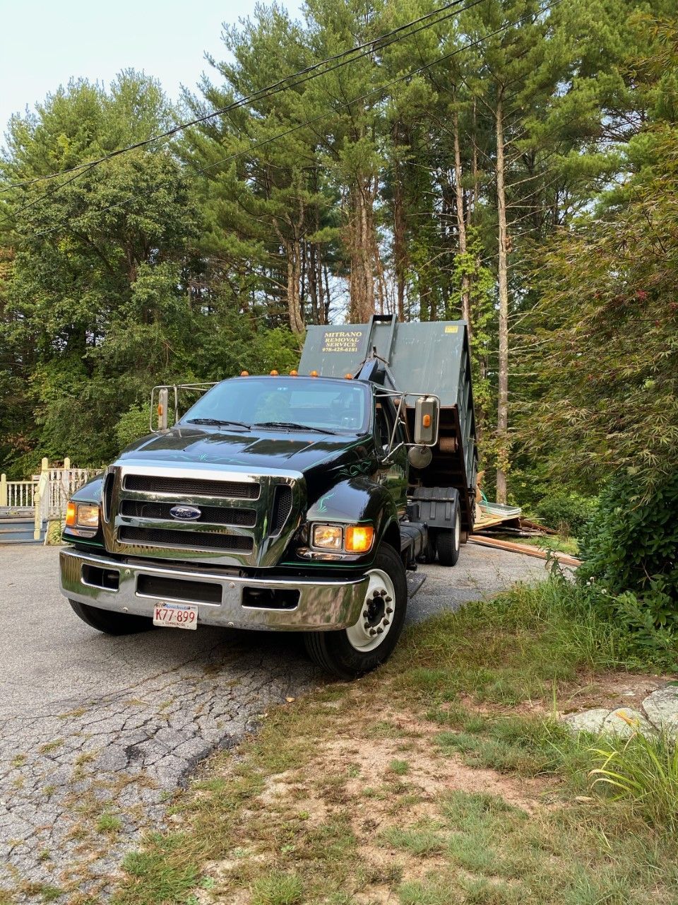 Black garbage truck on a gravel driveway, preparing to collect waste. Trees in the background.