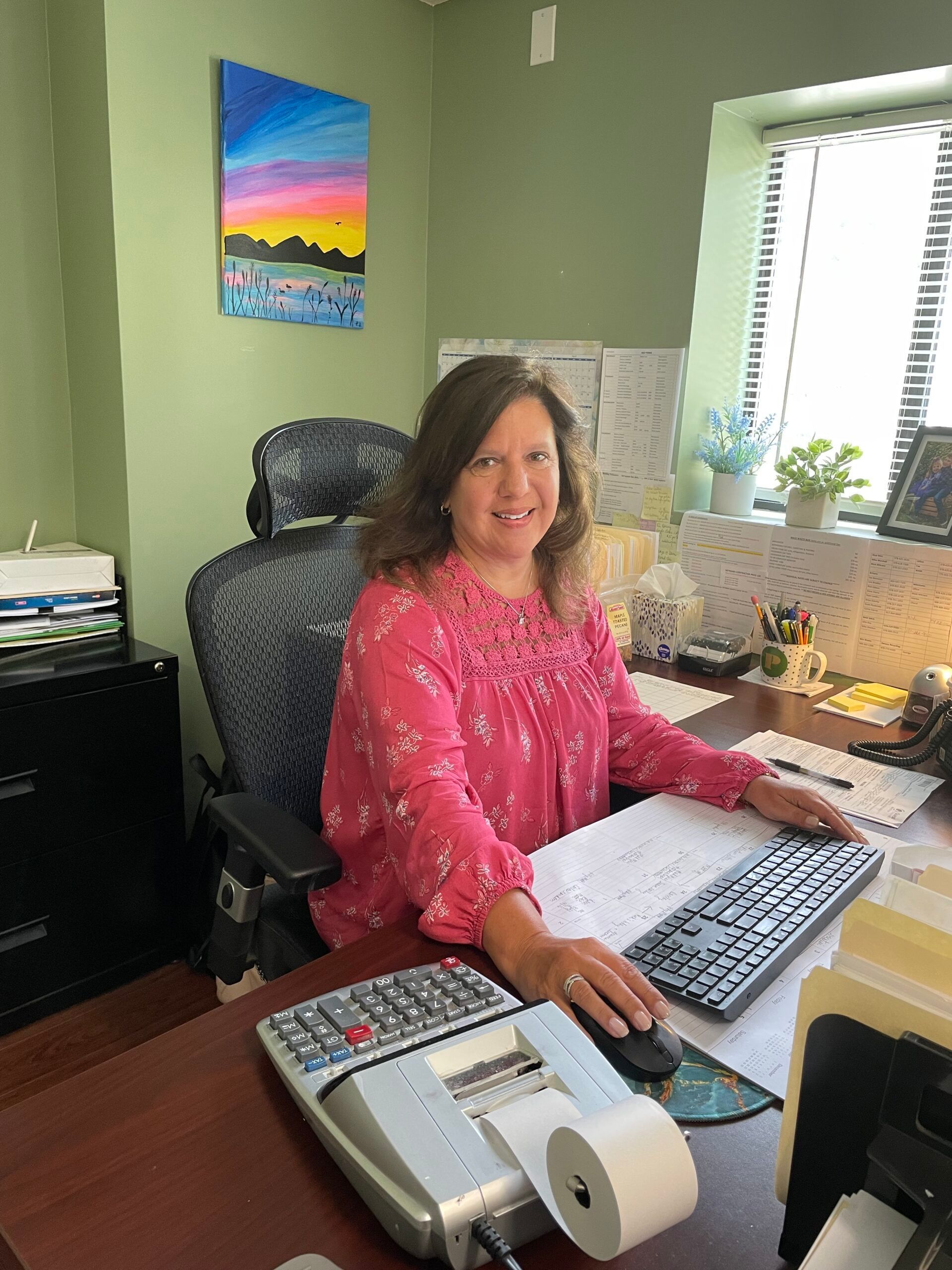 Woman in pink floral blouse at a desk, smiling. Office with a calculator, keyboard, and artwork.