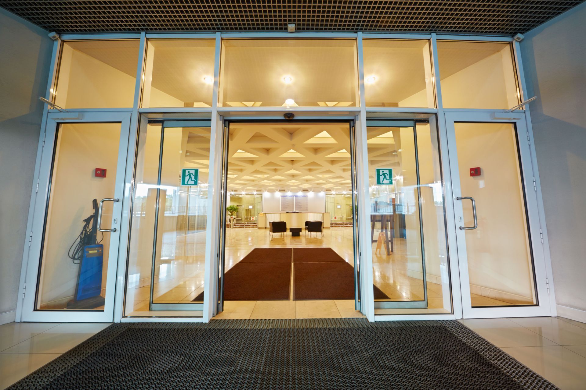 Glass entrance doors to a brightly lit building interior with a patterned ceiling and chairs.