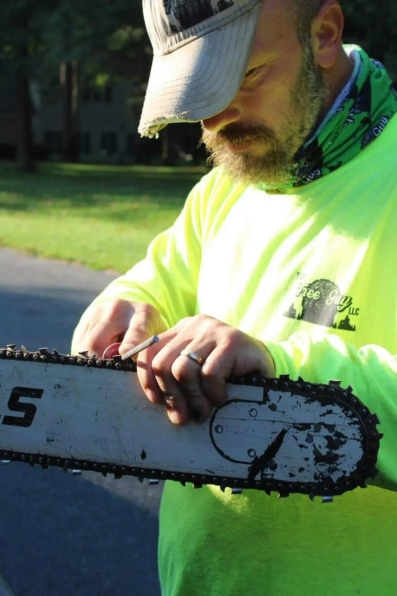 Man sharpening chainsaw outdoors, wearing a green long sleeve shirt and a hat.