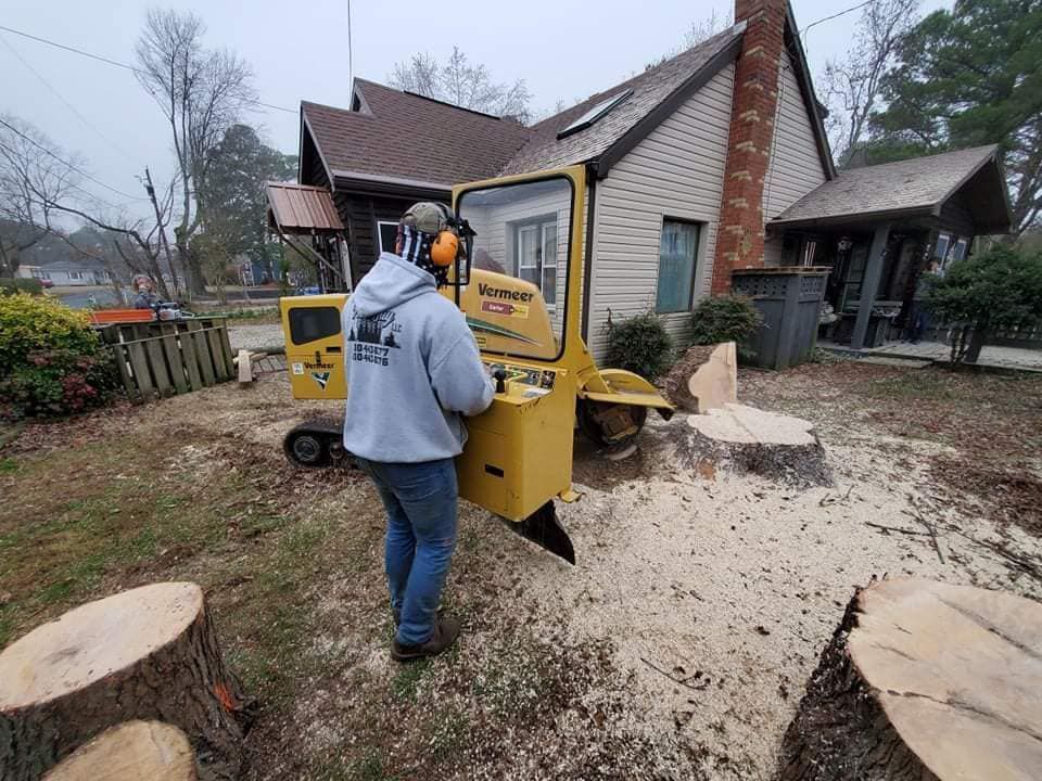 Man operating a yellow stump grinder near a house; wood chips on the ground.