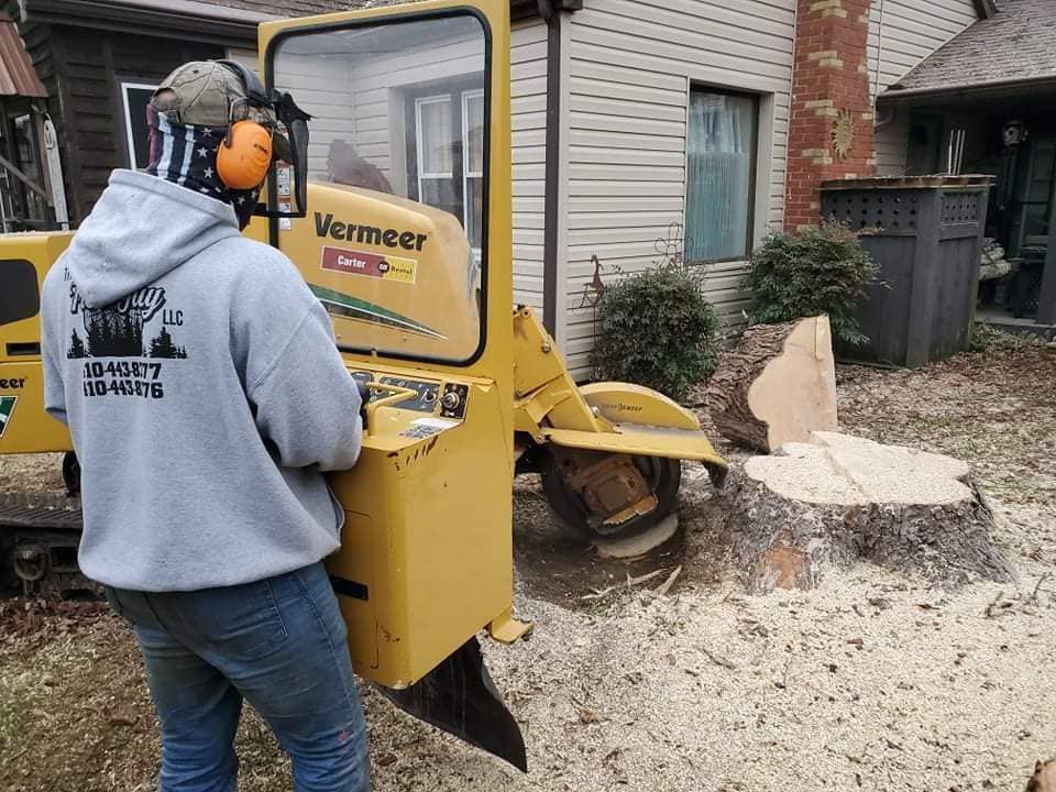 Man operating a yellow Vermeer stump grinder, grinding a tree stump in a yard, wearing hearing protection.