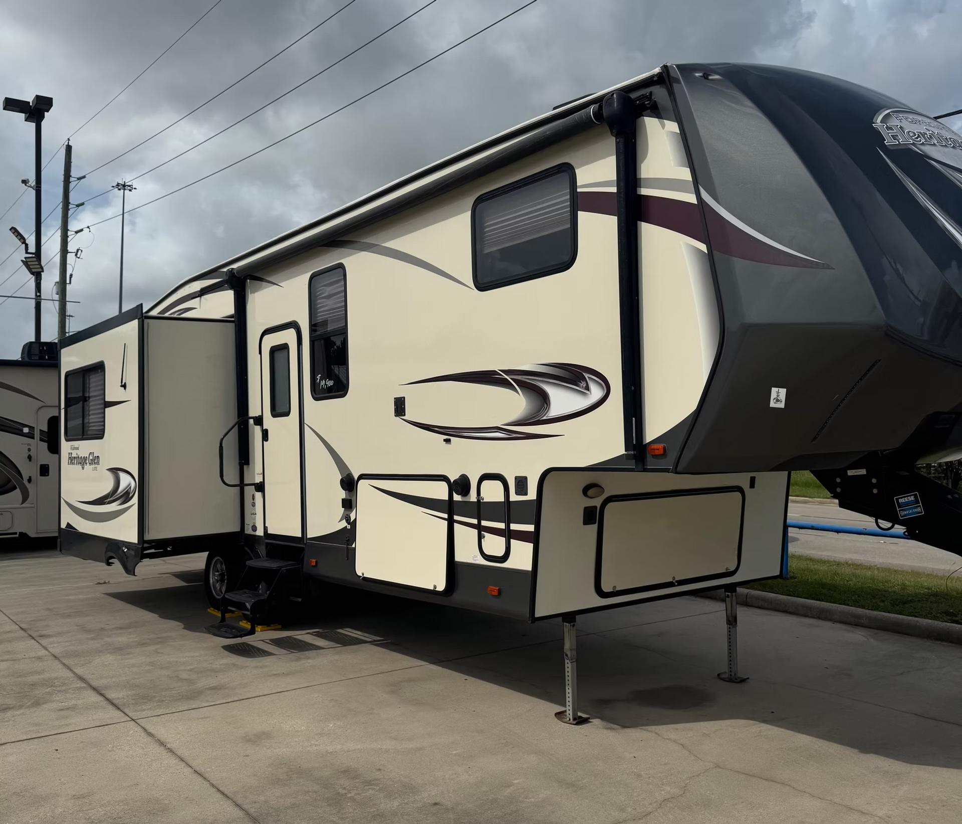 Large beige and black travel trailer parked on a concrete lot under a cloudy sky.