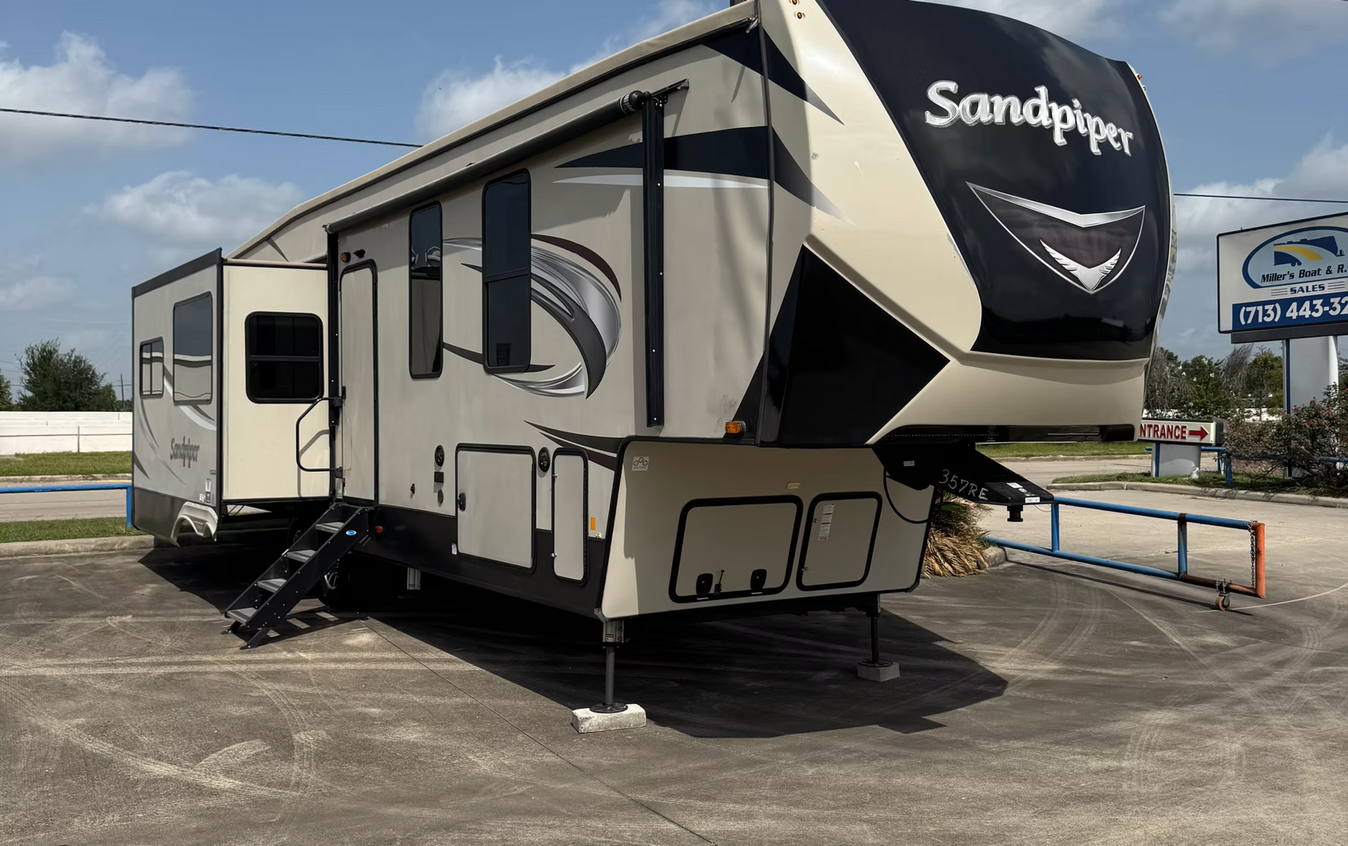 Black and beige Sandpiper fifth-wheel RV parked on pavement under a blue sky