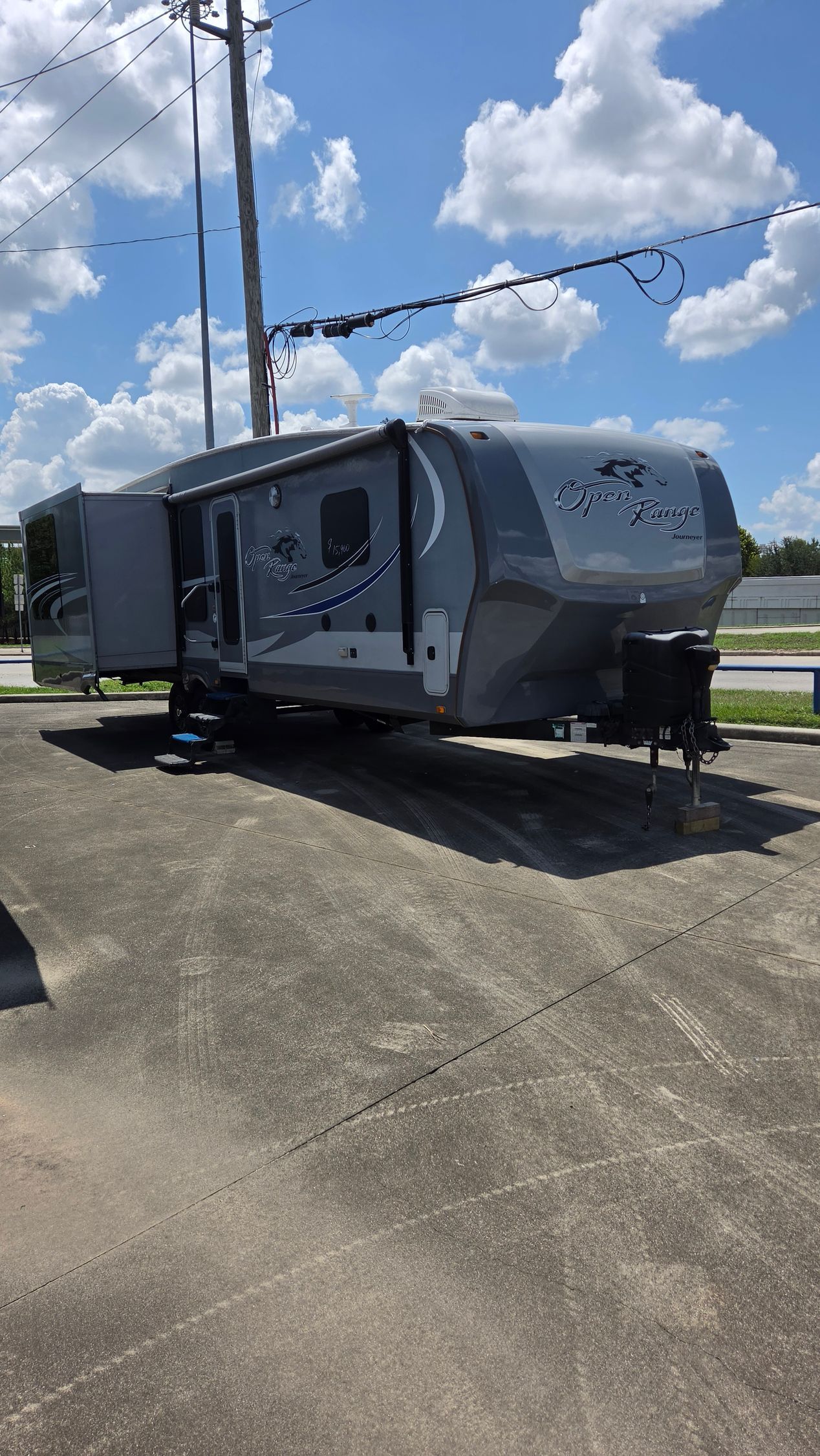 2015 Open Range 337 RLS - Gray travel trailer parked on a concrete lot under a blue sky with power lines and clouds.