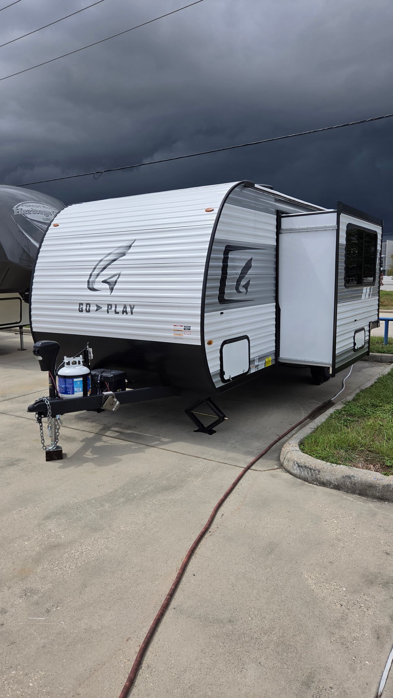 2016 Highland Go Play 18MBS - White travel trailer parked on a wet driveway under dark storm clouds, with a small awning and nearby chairs.