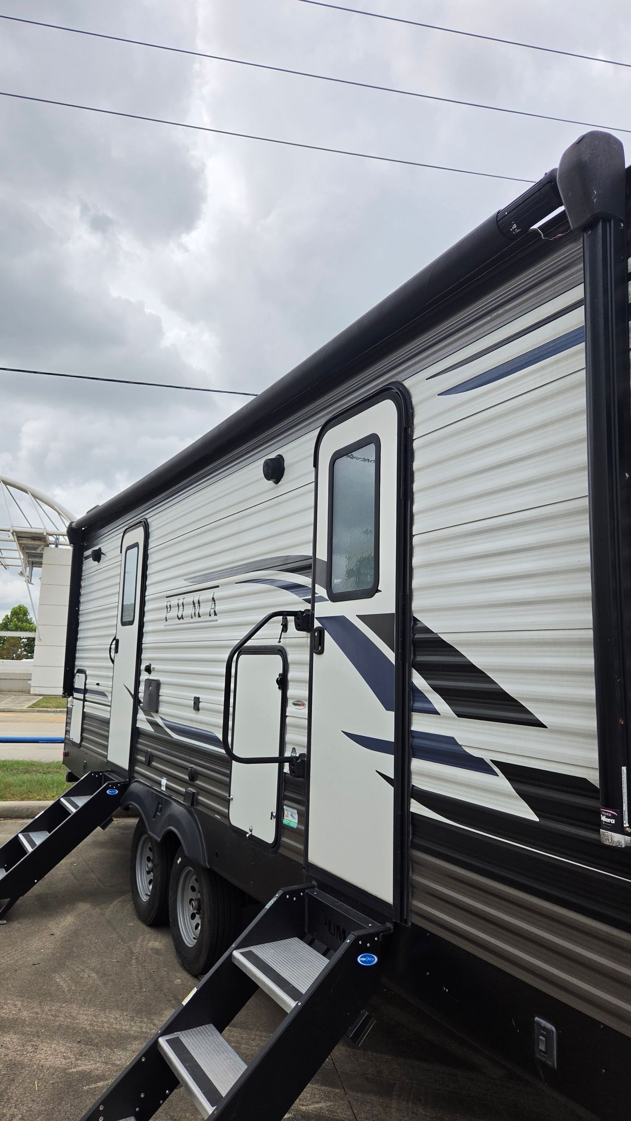 Black-and-white travel trailer with steps and awning parked on a gravel lot under cloudy sky