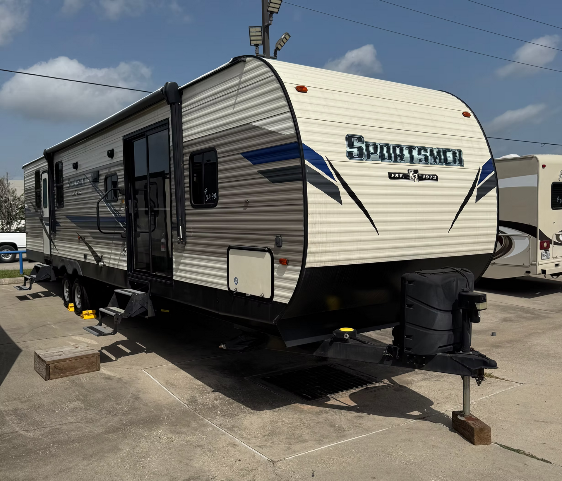White Sportsmen travel trailer parked on a lot, with blue graphics and rooftop equipment.