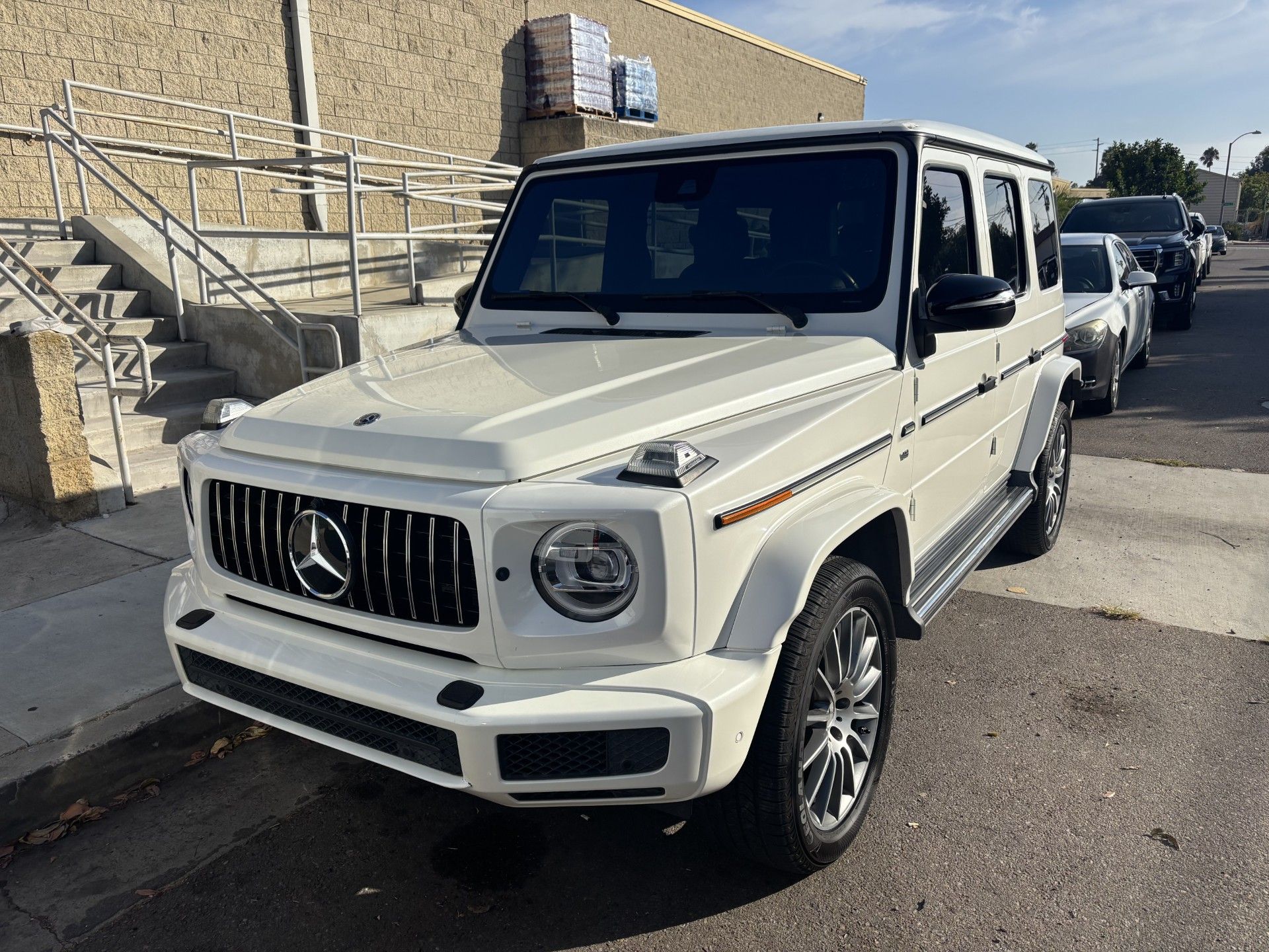 White Mercedes-Benz G-Class SUV parked beside a beige building with stairs and other cars in the lot