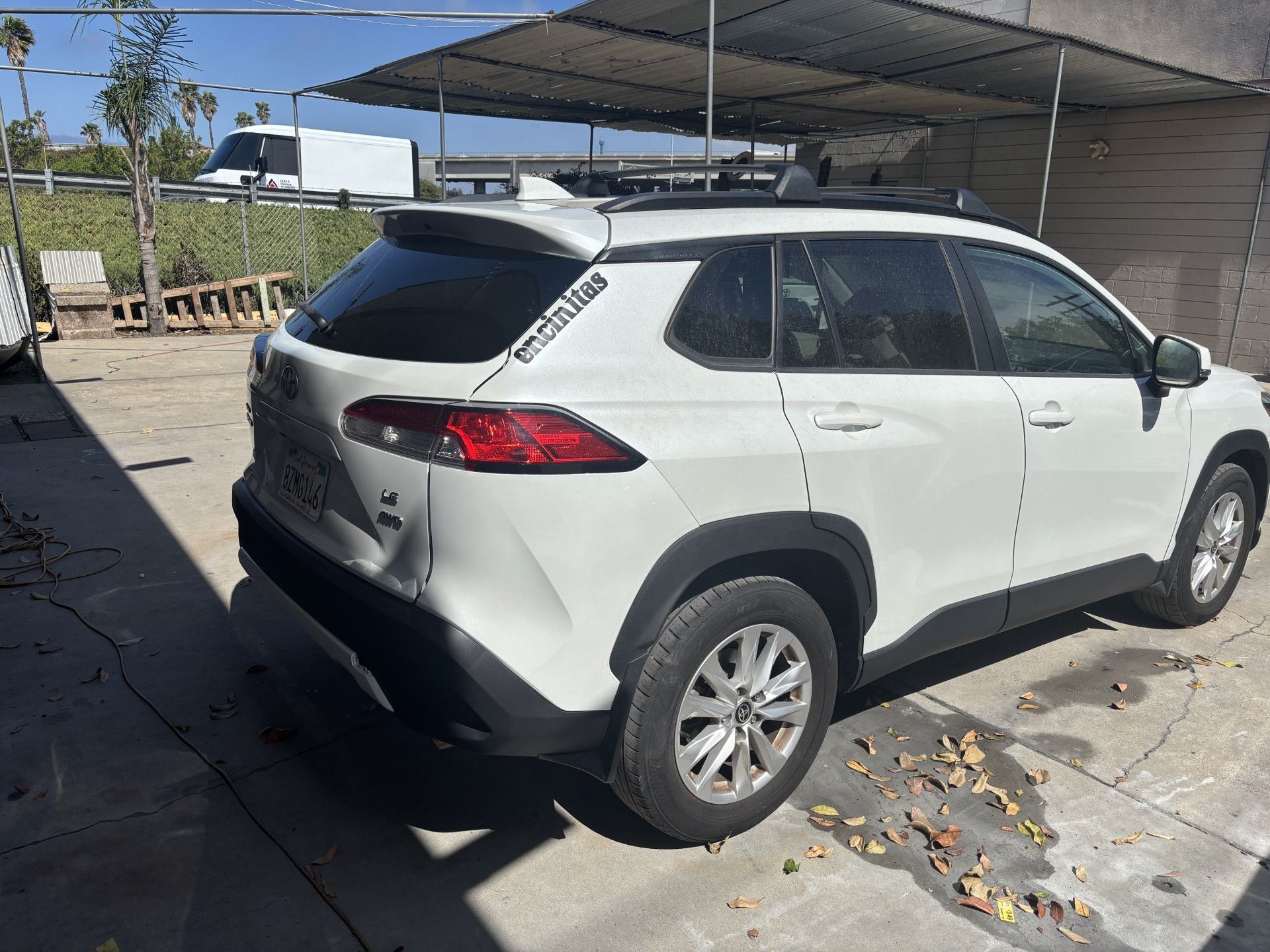 White SUV parked under a metal carport on a sunny paved lot, rear and side view.
