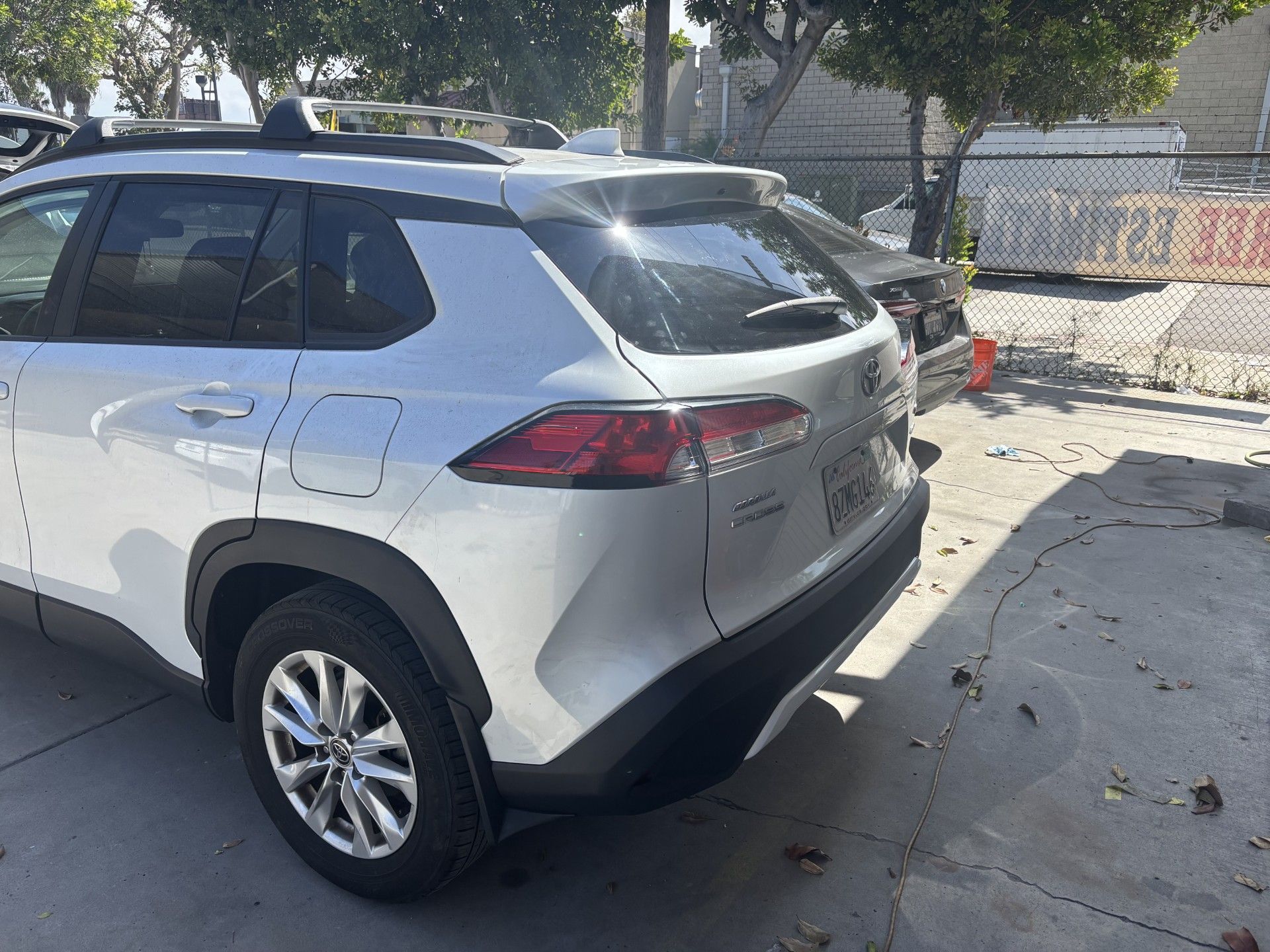 Silver SUV parked on a sunny street beside a sidewalk and chain-link fence.