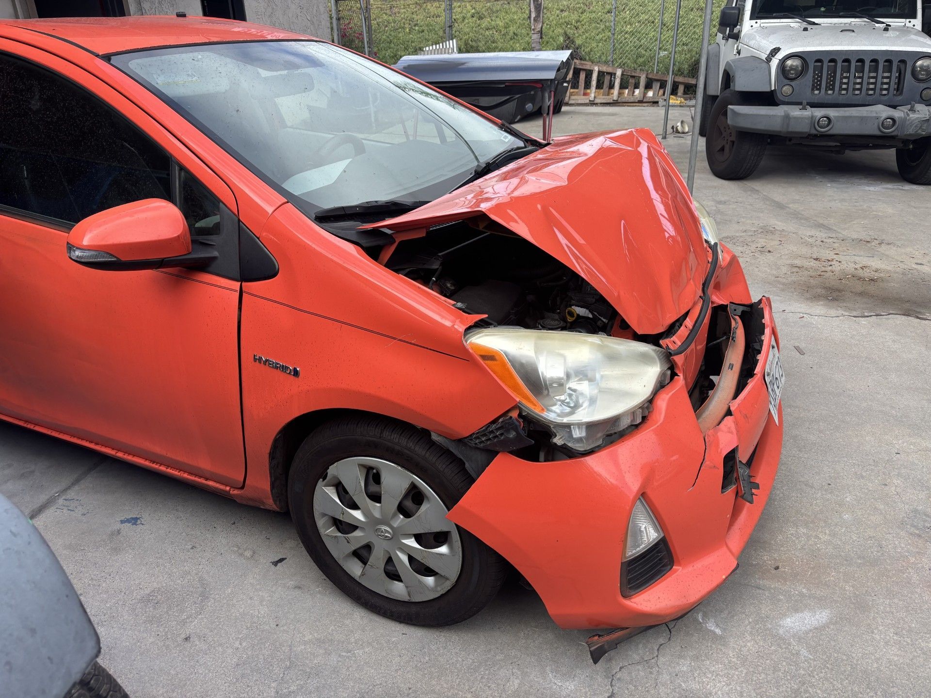 Red car with front-end collision damage, hood crumpled and bumper torn off, parked beside a Jeep.