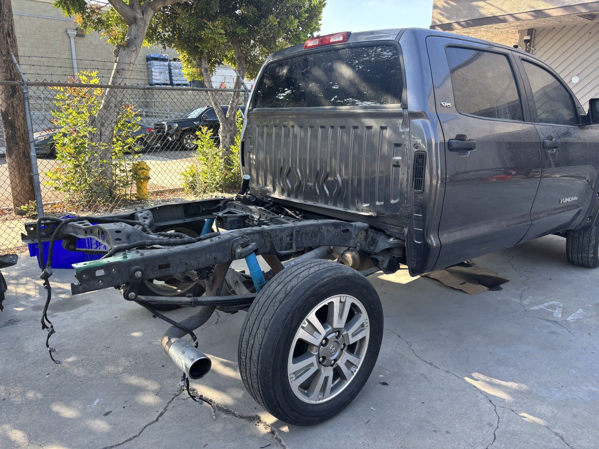 Damaged black pickup truck with missing bed, parked on a driveway near a fence and trees