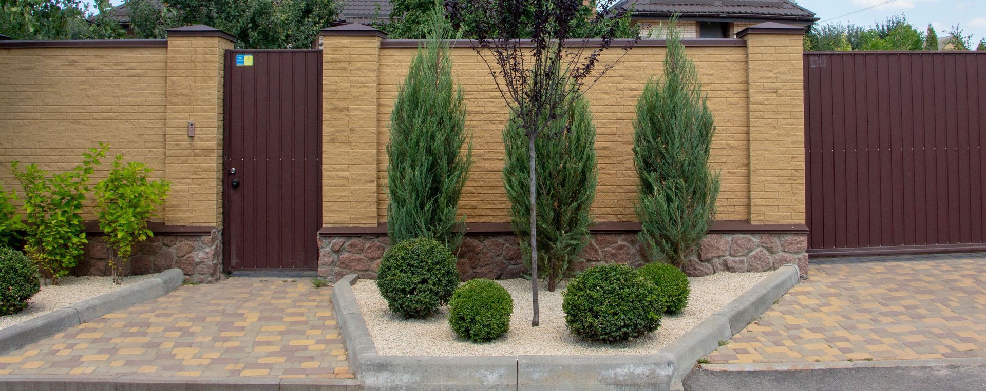 A residential garden bed featuring three tall, slim evergreen trees and four small, rounded bushes against a tan wall.