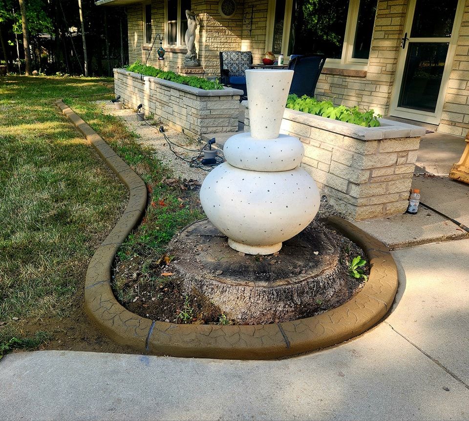 A white, three-tiered spherical fountain sits in a circular, concrete-bordered garden bed next to a stone-faced house.