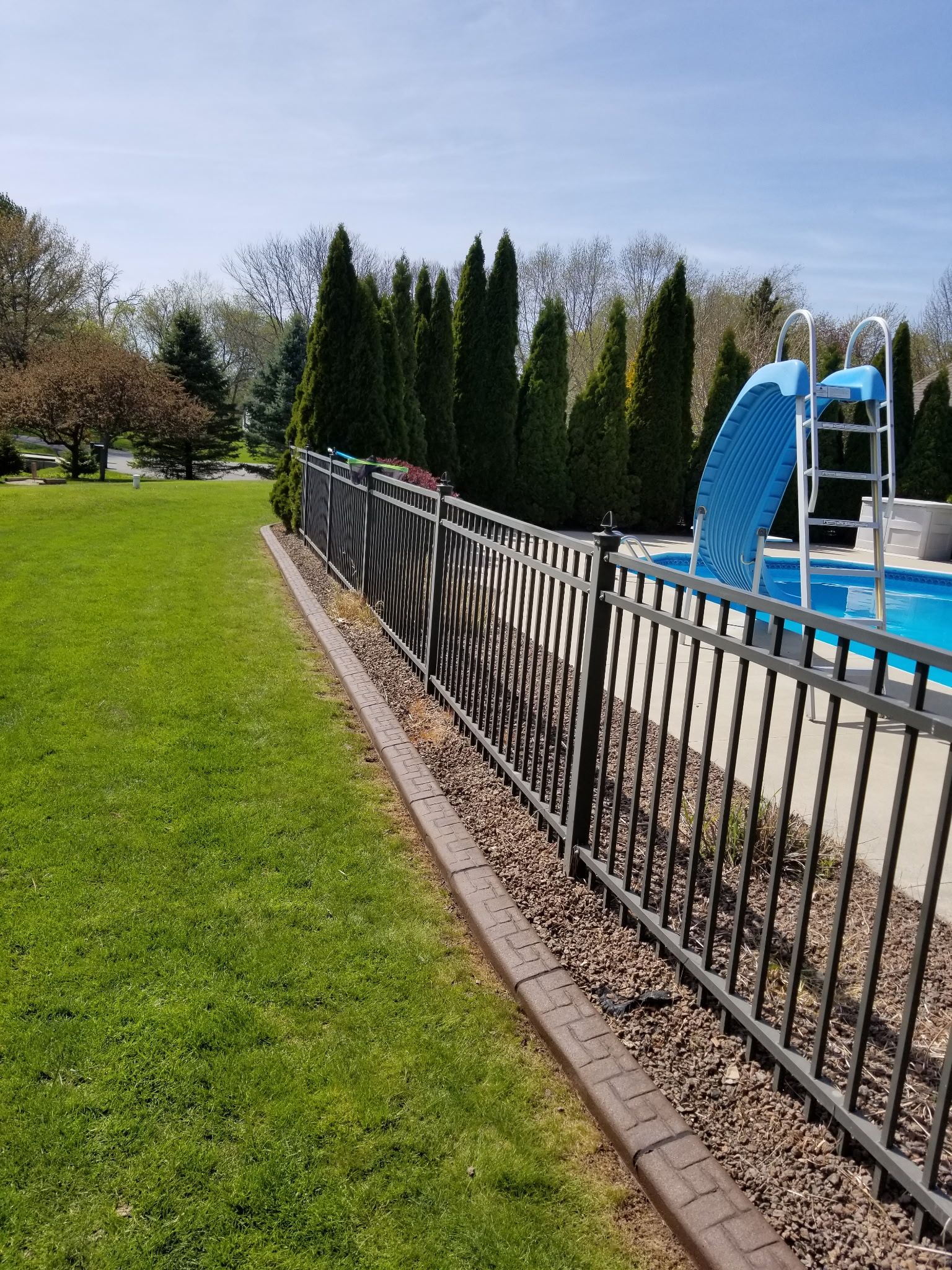 Backyard pool with blue slide beside a black fence and green lawn on a sunny day