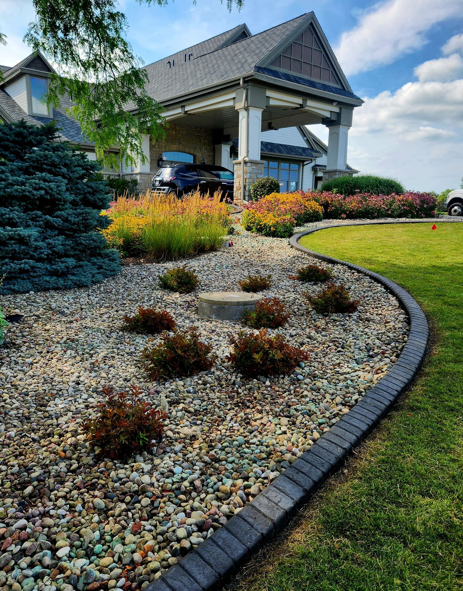 Landscaped front yard with colorful flower beds, gravel mulch, and a house porch under a blue sky