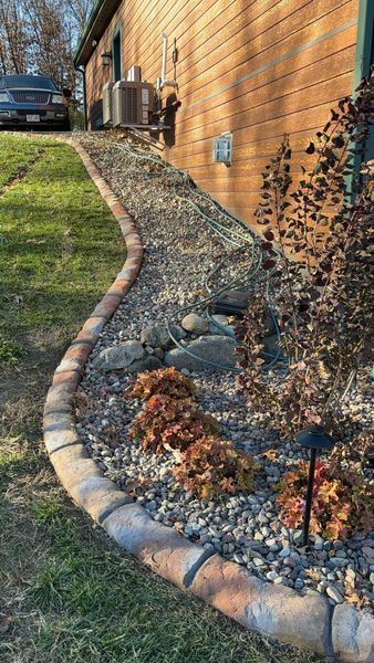 A landscape bed along a wood-sided house, featuring grey gravel, small shrubs, stone edging, and a curving lawn border.