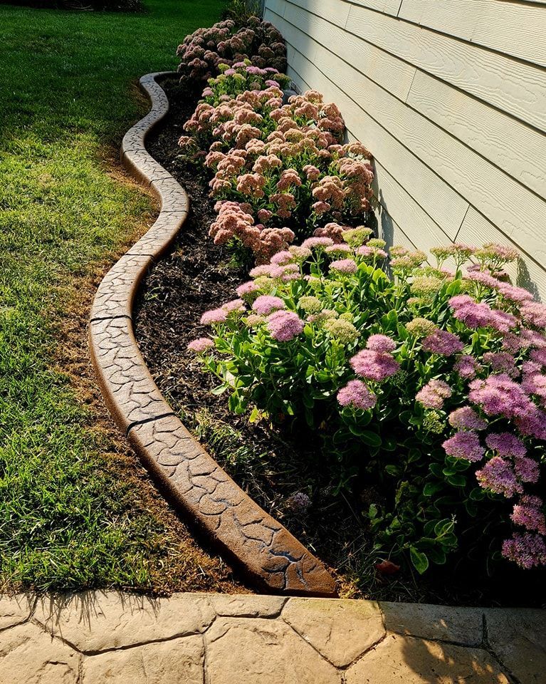 A curved, stamped concrete border separates a mulch garden bed with pink sedum flowers from a grassy lawn.