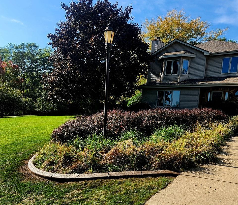 A decorative landscape bed with a lamppost, dark purple shrubs, and ornamental grasses beside a grey house.