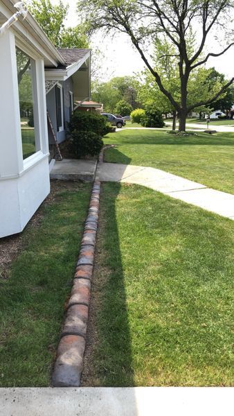 A row of decorative stone pavers separates a lawn from a house exterior, leading to a sidewalk in a residential yard.