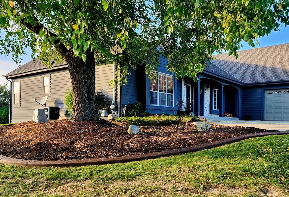 A blue house with a large tree in the front yard featuring a curved, mulched garden bed bordered by landscape stones.