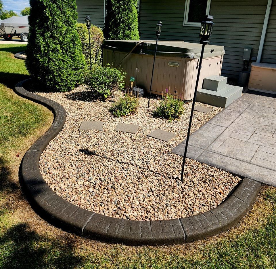 A curved dark stone border frames a gravel landscaping area featuring small shrubs and a hot tub on a concrete patio.