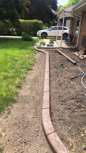 A curved, brown textured concrete landscape edging installed in a yard between a grass lawn and a dirt flower bed area.
