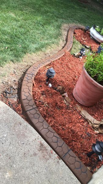 Curved, decorative stamped concrete landscape edging separating red mulch from grass, next to a concrete patio.