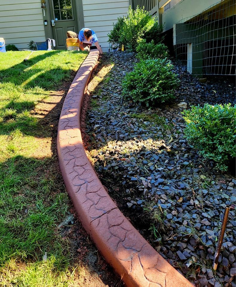 A person installs a decorative, reddish-brown stamped concrete border between a green lawn and a rock-filled garden bed.