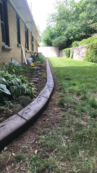 A garden bed edged with timber ties runs along the side of a tan brick house, separating it from a grass lawn.