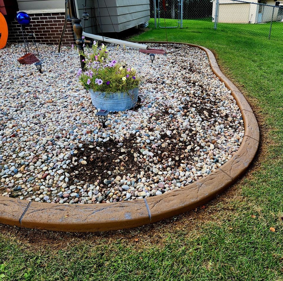 A curved, stamped concrete border edges a gravel garden bed containing a potted plant on a grassy lawn.