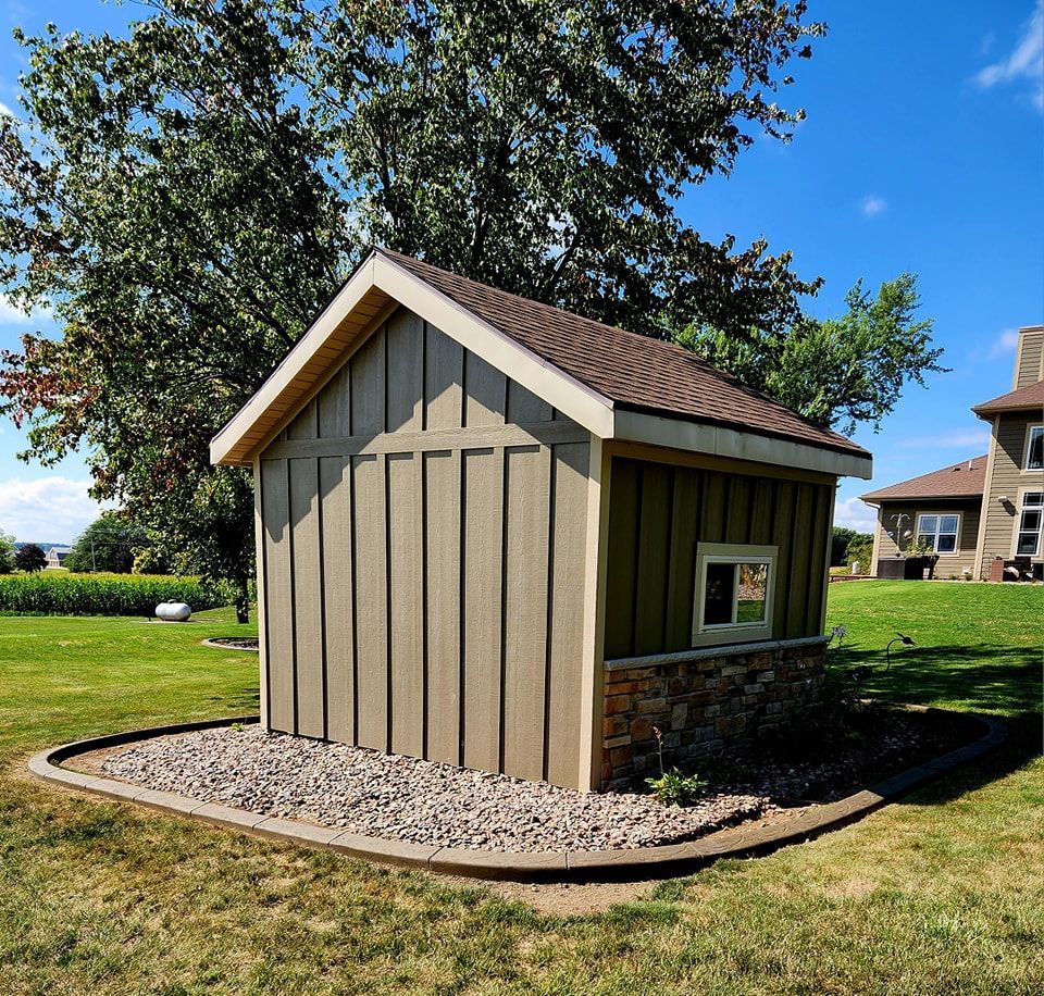 A small, tan storage shed with a brown shingled roof, a small window, and a stone base, surrounded by gravel on a lawn.