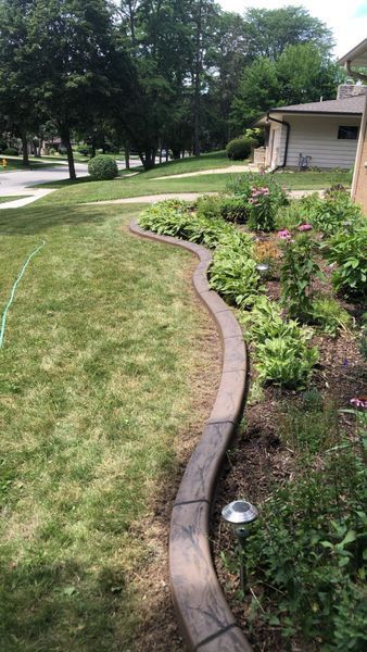 A curving concrete landscape border separates a green lawn from a garden bed with mulch, plants, and solar lights.
