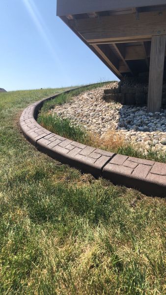 A curved, brown, stamped concrete landscape border separates a grassy lawn from a gravel-filled area under a deck.