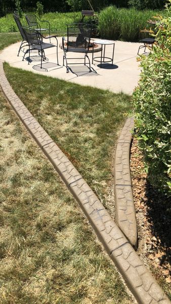 A stone-textured landscape border curves through a grassy lawn leading toward a patio with chairs and a fire pit.