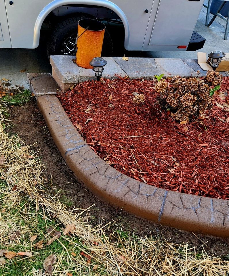 A curved, stamped concrete landscape border surrounds a mulch bed with a dry plant and solar lights near a parked trailer.