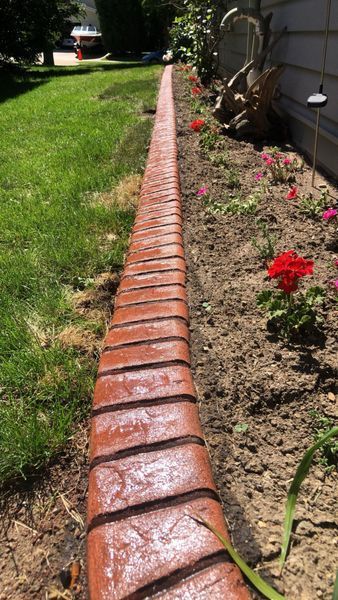 A curved, red brick-patterned landscape border separates a grassy lawn from a flower garden near a house wall.