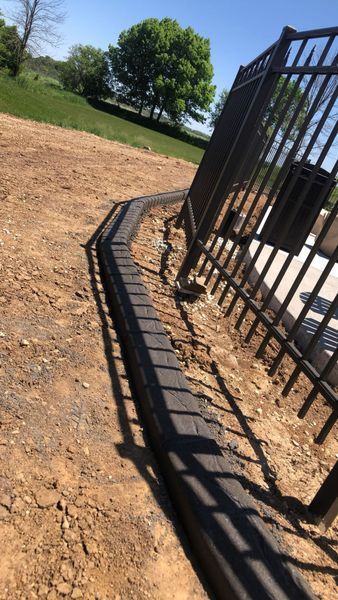 A dark metal fence stands next to a recently installed row of dark concrete landscaping bricks on a dirt path.