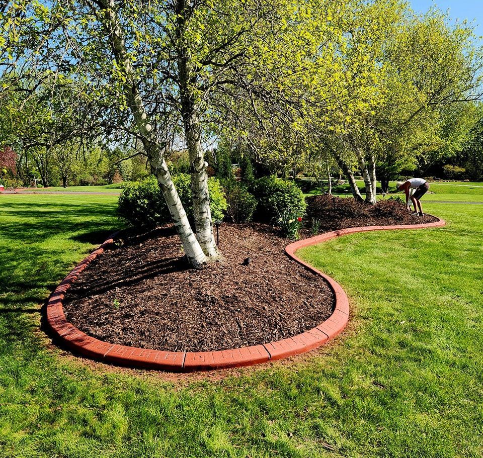 A gardener tends to a mulched tree bed bordered by curved red bricks in a lush, green lawn under a sunny sky.