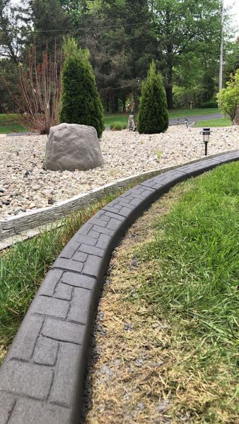 Decorative gray concrete landscape edging separates a manicured green lawn from a rock-filled garden bed with evergreens.