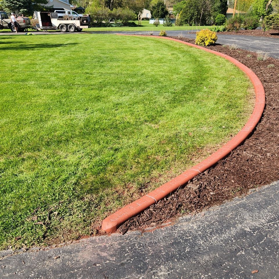 A curved, reddish-brown concrete landscape border separates a lush green lawn from a dark asphalt driveway and mulch bed.