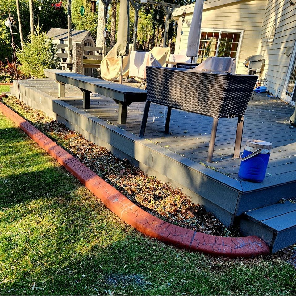 A backyard deck with a dark woven bench and table, featuring a red concrete landscape border along a grassy lawn.