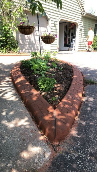 A triangular brick garden bed sits in a driveway, with two hanging baskets above and a person walking toward a garage.