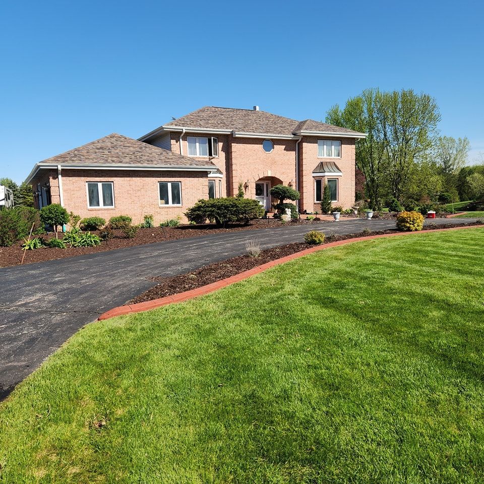 A two-story brick house with a large paved driveway and green lawn under a clear blue sky.