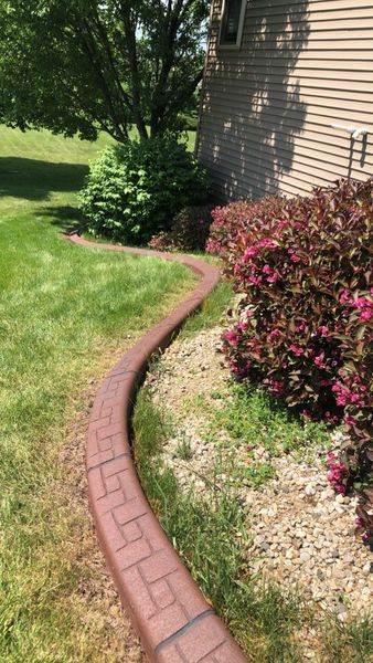 A winding, brick-patterned concrete landscape border separates a grassy lawn from a mulched flower bed beside a house.